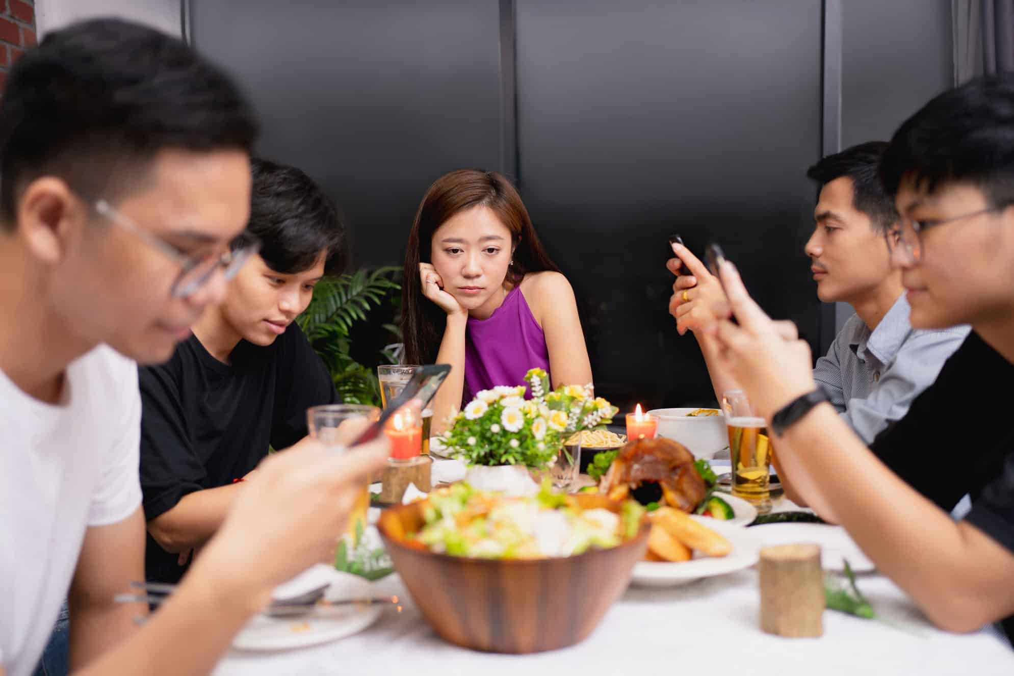Family on a dinner table filled with food, one woman looks sad, everyone else are on their phones