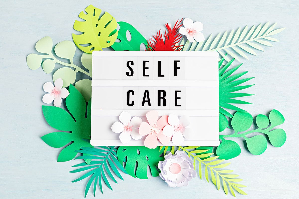 The words "self care" on a board with flowers and leaves in the background.