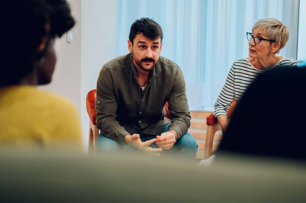 Man seated around a group of people, talking.