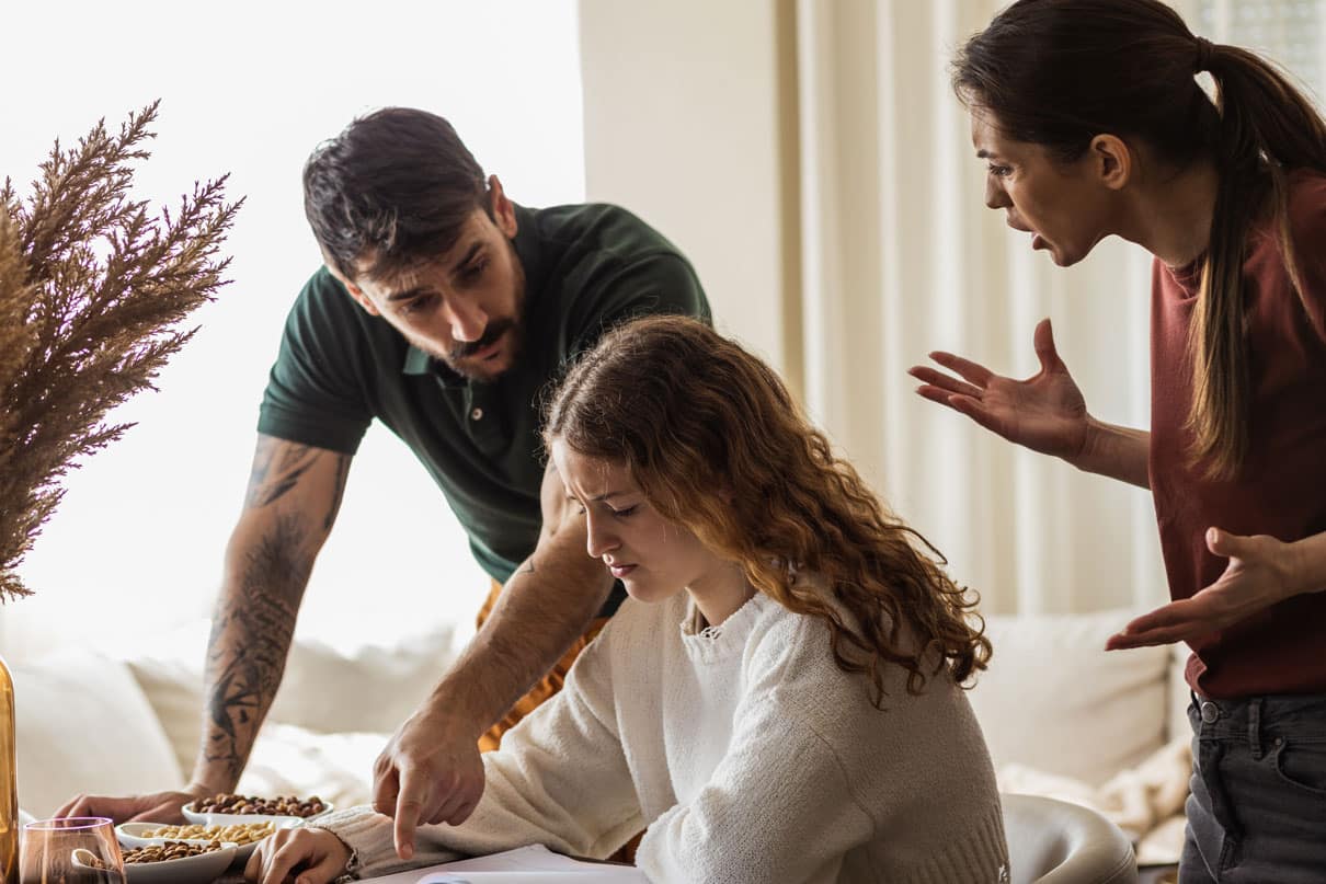 Teenage girl sitting down, looking visibly distressed, surrounded her parents talking to her, looking upset.